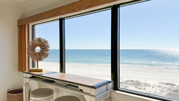Una habitación con un ventanal grande que da a la playa, una mesa blanca con dos taburetes y una cesta tejida en el suelo.