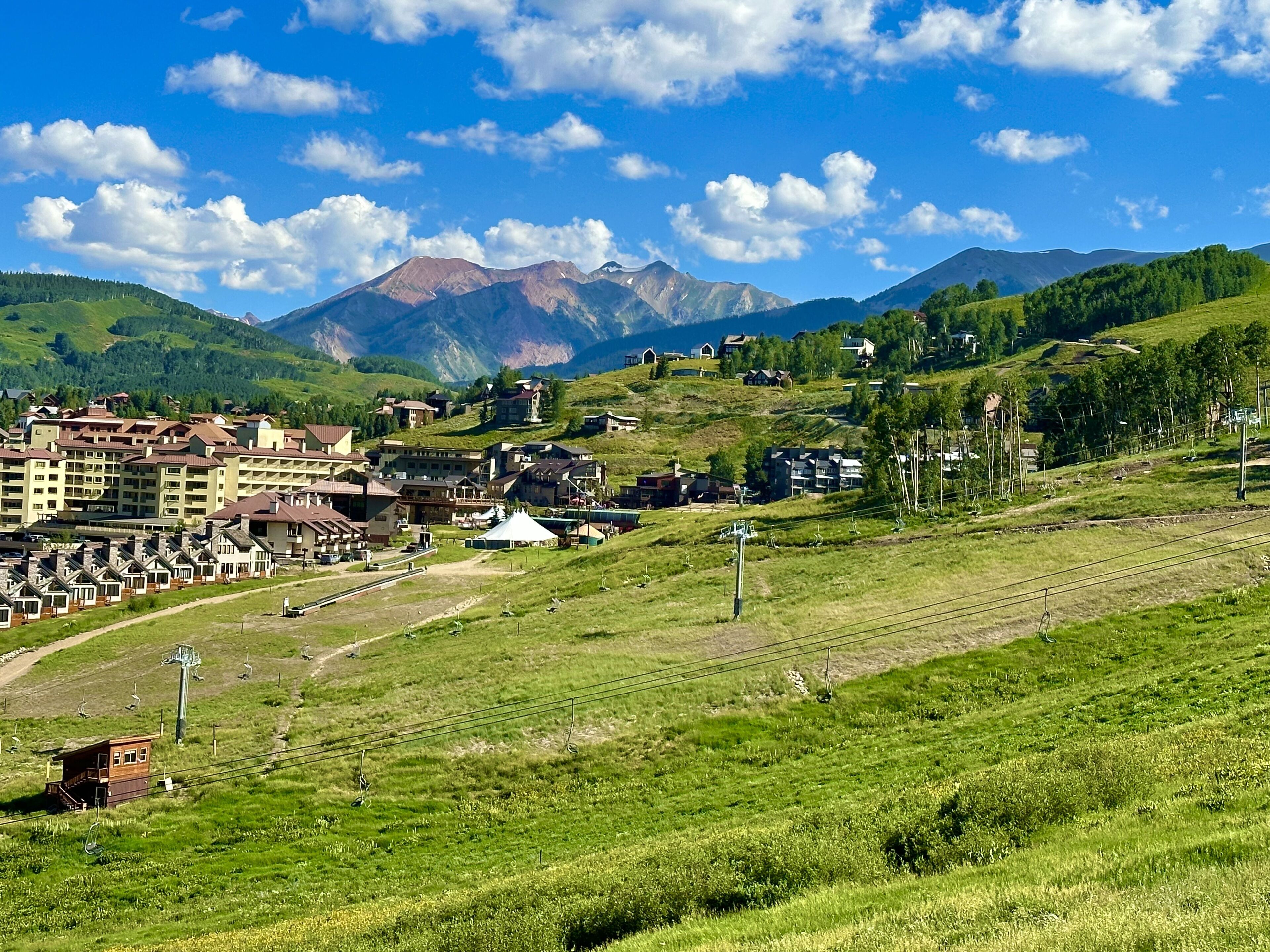 A mountainous landscape with a town featuring multi-story buildings, a ski lift, and a clear blue sky.