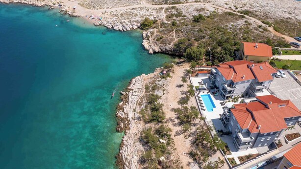 An aerial view of a coastal area with a rocky shoreline, clear turquoise waters, and a resort with red-tiled buildings and a swimming pool.