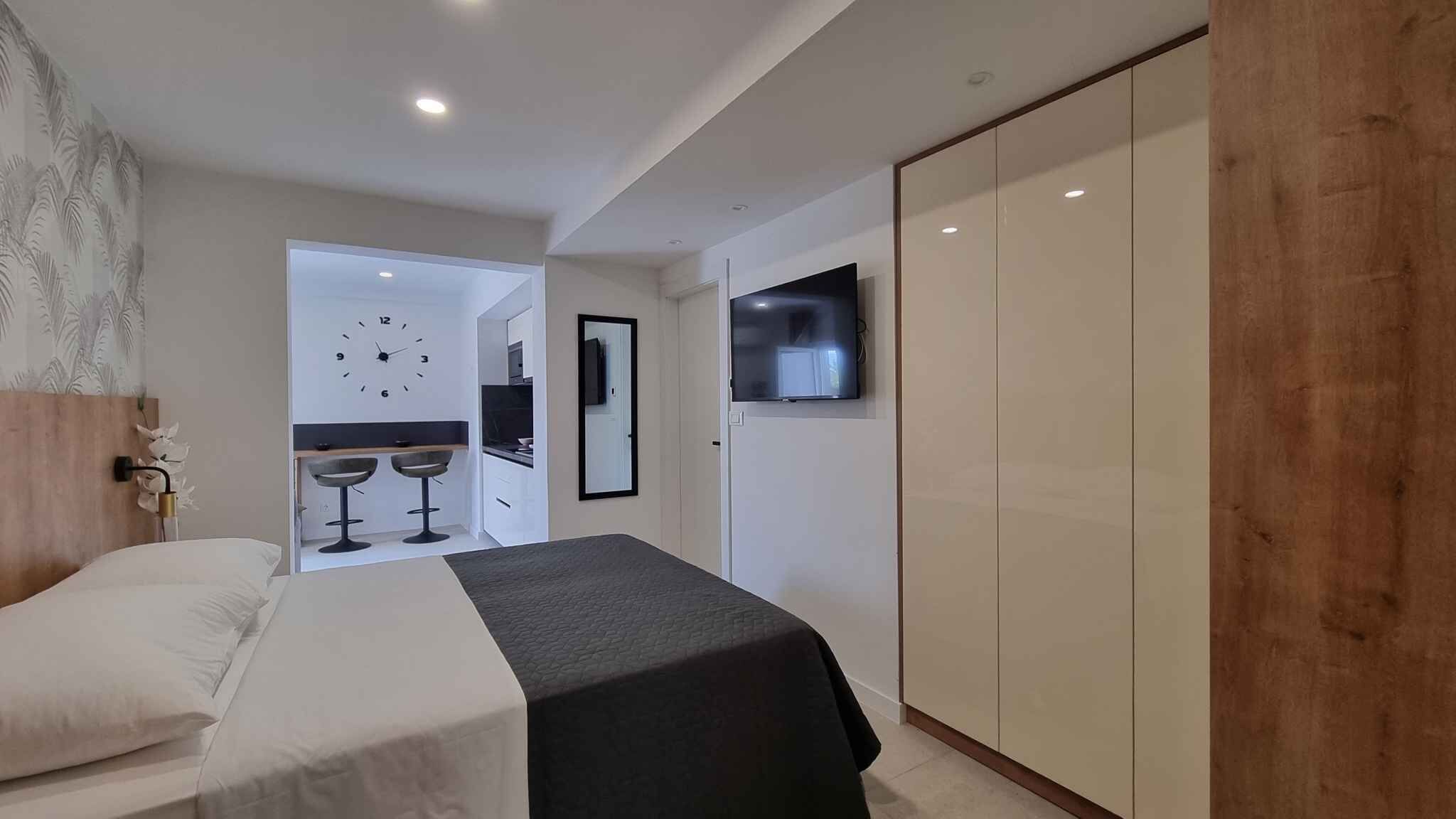 A modern hotel room with a bed, a TV mounted on the wall, and a separate dining area with two bar stools.