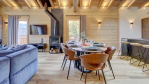 Een moderne woonkamer met een open haard, houten balken in het plafond, een eethoek met een tafel voor vier personen en een aan de muur bevestigde televisie.