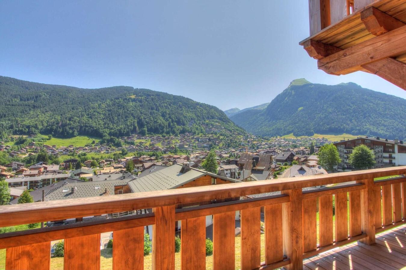 A balcony with a wooden railing overlooking a village nestled in a valley with mountains in the background.