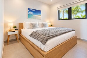 A modern bedroom with a wooden bed, white bedding, a grey throw, a nightstand with a lamp, a clock, and a framed picture of a beach. - 25% Off Oct 15-18 Pool, Grace Bay, Ocean View Z102 (Grace Bay)