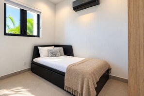 A bedroom with a bed, a window with a view of palm trees, and a wall-mounted air conditioner.