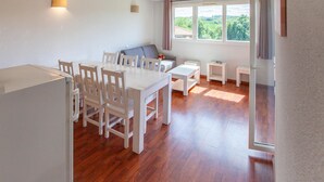 Un vaste espace de vie comprenant une table à manger, des chaises, un canapé et une petite table basse. Une fenêtre avec des rideaux, une lampe et une vue sur la verdure à l’extérieur complètent l’ensemble.