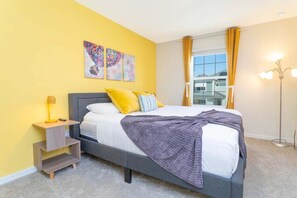 Ein Schlafzimmer mit Bett, Nachttisch, Lampe, einem Fenster mit Vorhängen und einem Bild an der Wand.