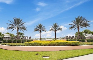 Entrada de un resort con palmeras, cielo despejado y césped cuidado.
