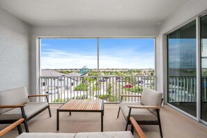 A balcony with two chairs and a table, a clear view of a parking lot and buildings, and a large glass door.