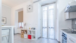 Une cuisine moderne équipée d’un four encastré, d’un micro-ondes et d’un évier. On y trouve une étagère blanche où sont posés des livres et des objets décoratifs. Une horloge est visible au mur.