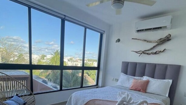 Ein Schlafzimmer mit großem Fenster, einem Bett mit weißer und rosa Decke, einem Deckenventilator und einem Sessel.