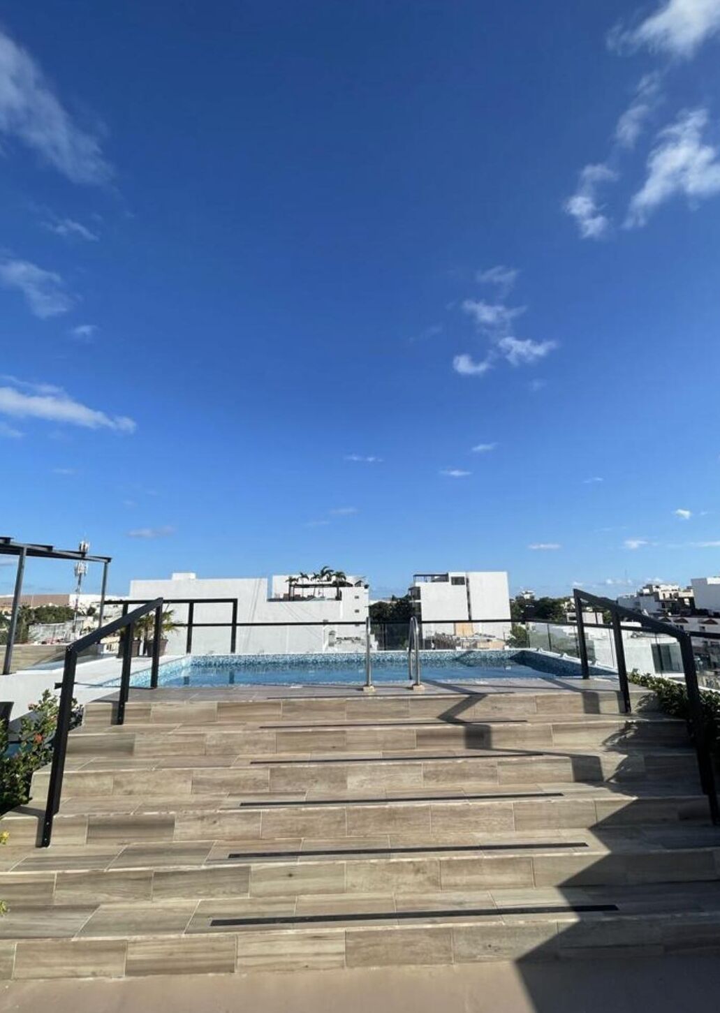 Ein Pool auf dem Dach mit Stufen, umgeben von modernen Gebäuden und blauem Himmel.
