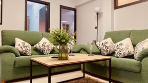 A green sofa with leaf-patterned pillows, a wooden coffee table, and a vase with green foliage.