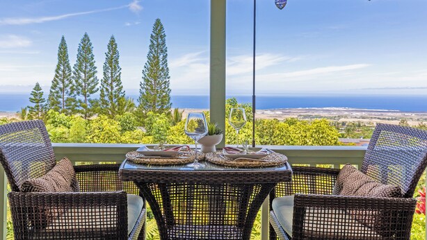 A covered outdoor seating area with wicker chairs and a table, overlooking a scenic view of trees and the ocean.