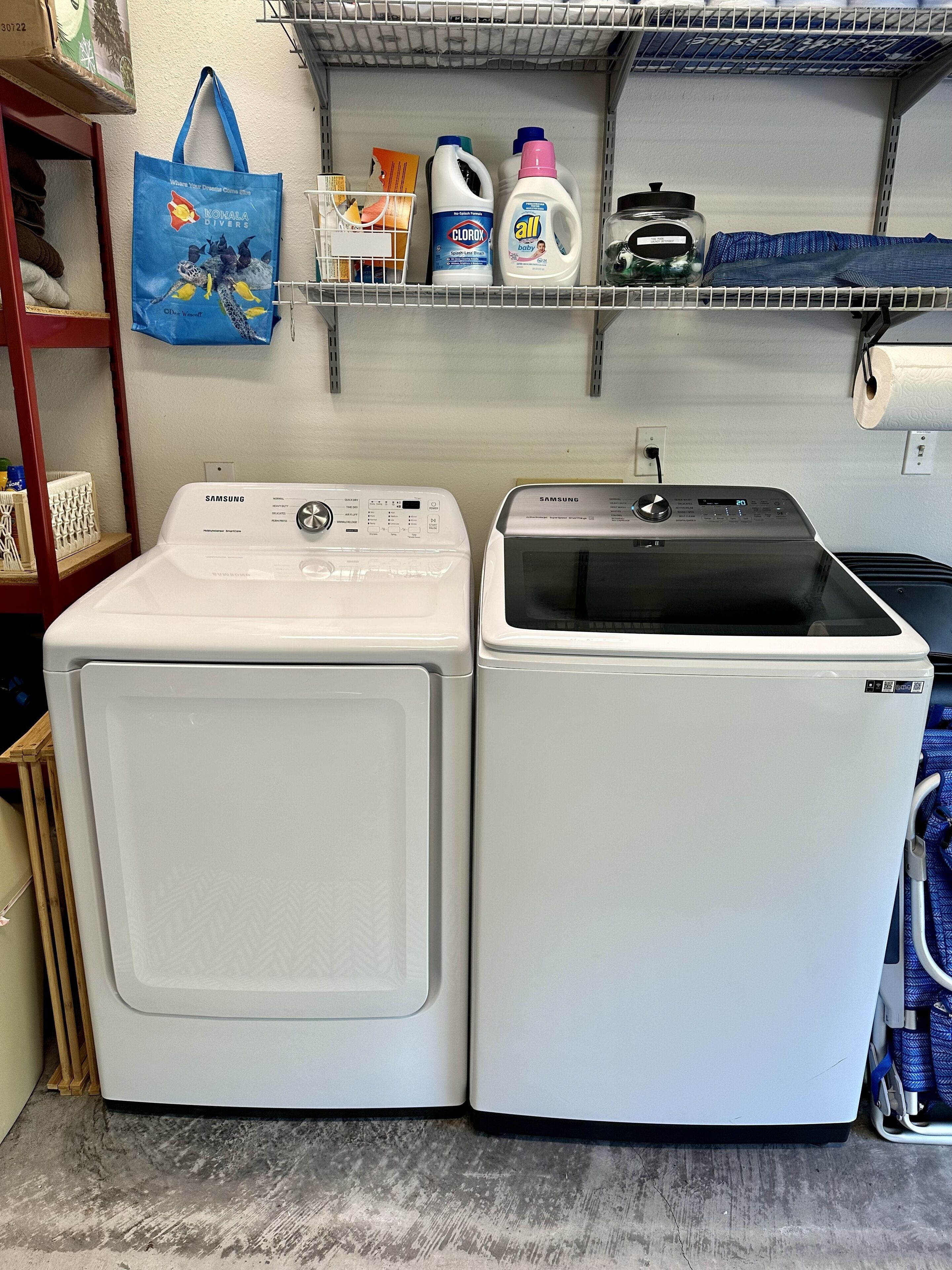 Two white Samsung washing machines side by side in a laundry room with shelves above containing cleaning supplies and a blue tote bag.