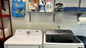Two white Samsung washing machines side by side in a laundry room with shelves above containing cleaning supplies and a blue tote bag. - Kailua-Kona 3-bedroom, 2-bath single-family home Ocean & Sunset View (Kailua-Kona)