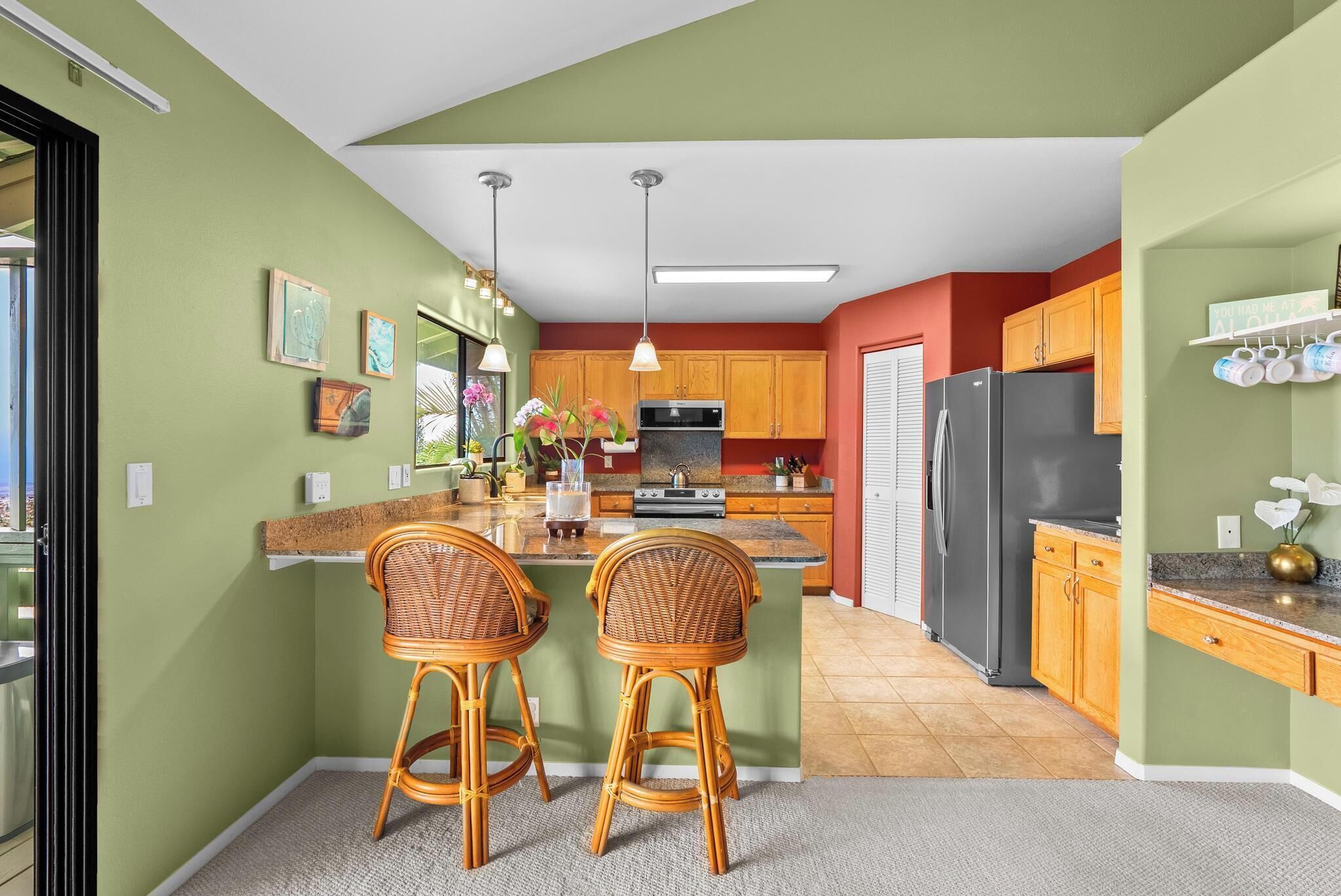 A kitchen with a central island, two wicker bar stools, wooden cabinets, a refrigerator, and a microwave.