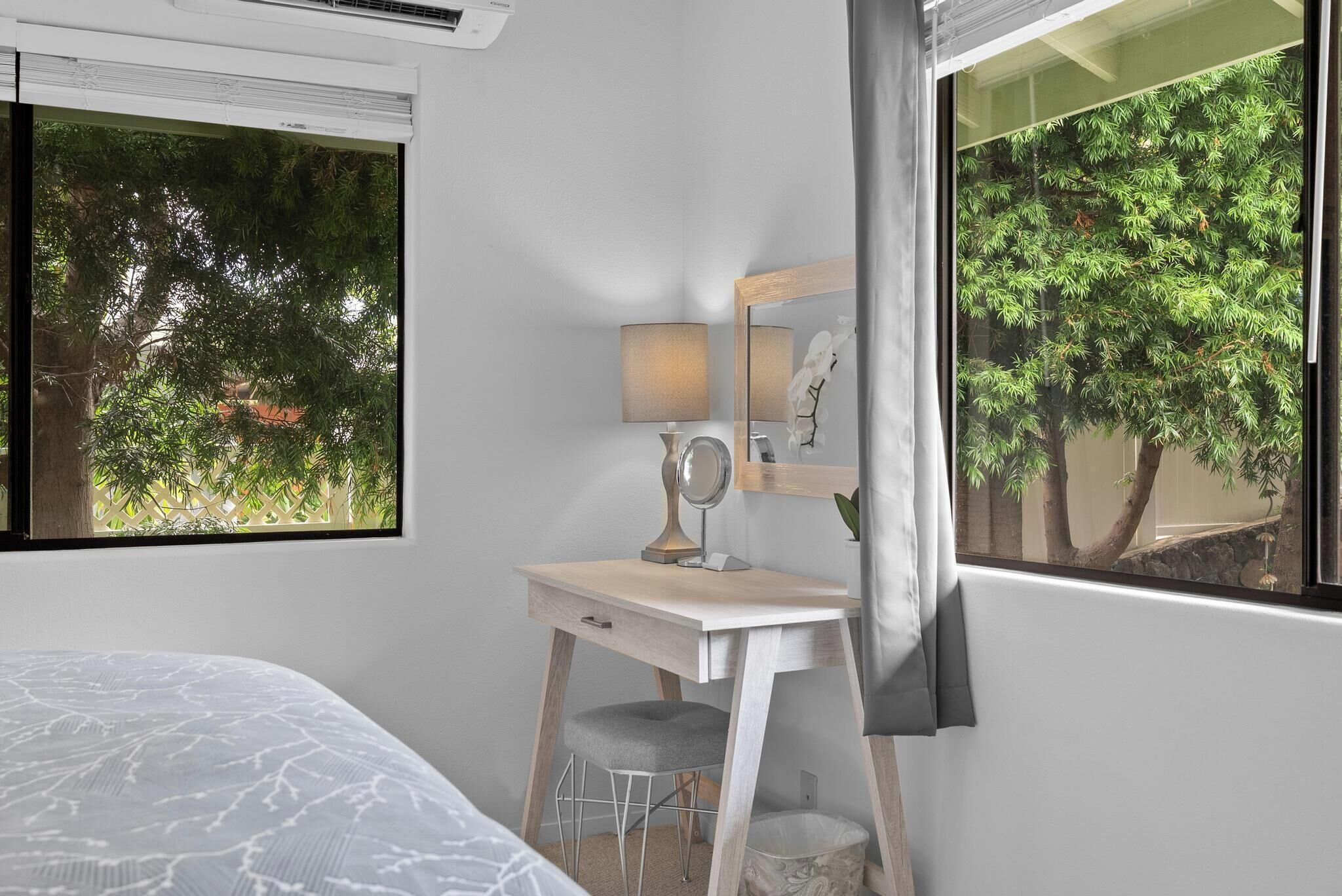 A bedroom with a bed, a desk with a mirror, a lamp, and a view of trees through the window.
