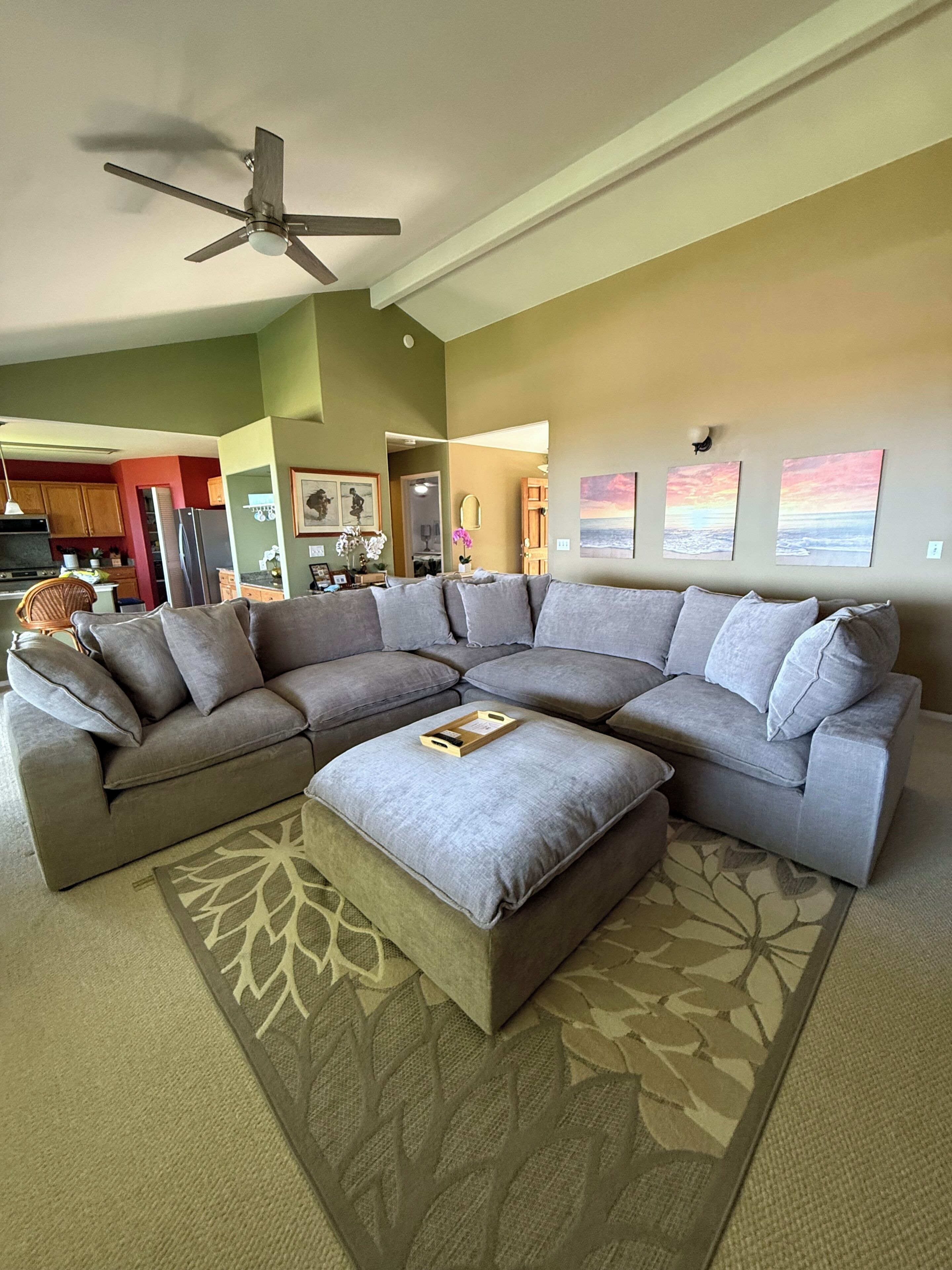 A living room with a sectional sofa, a ceiling fan, and a kitchen in the background.
