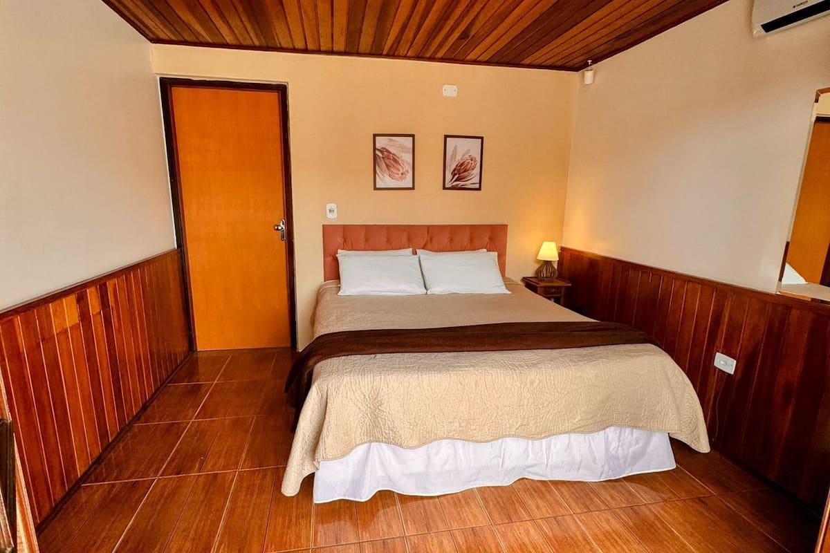 A hotel room with a wooden ceiling, a bed with a beige and white bedspread, two framed pictures on the wall, and a wooden floor.