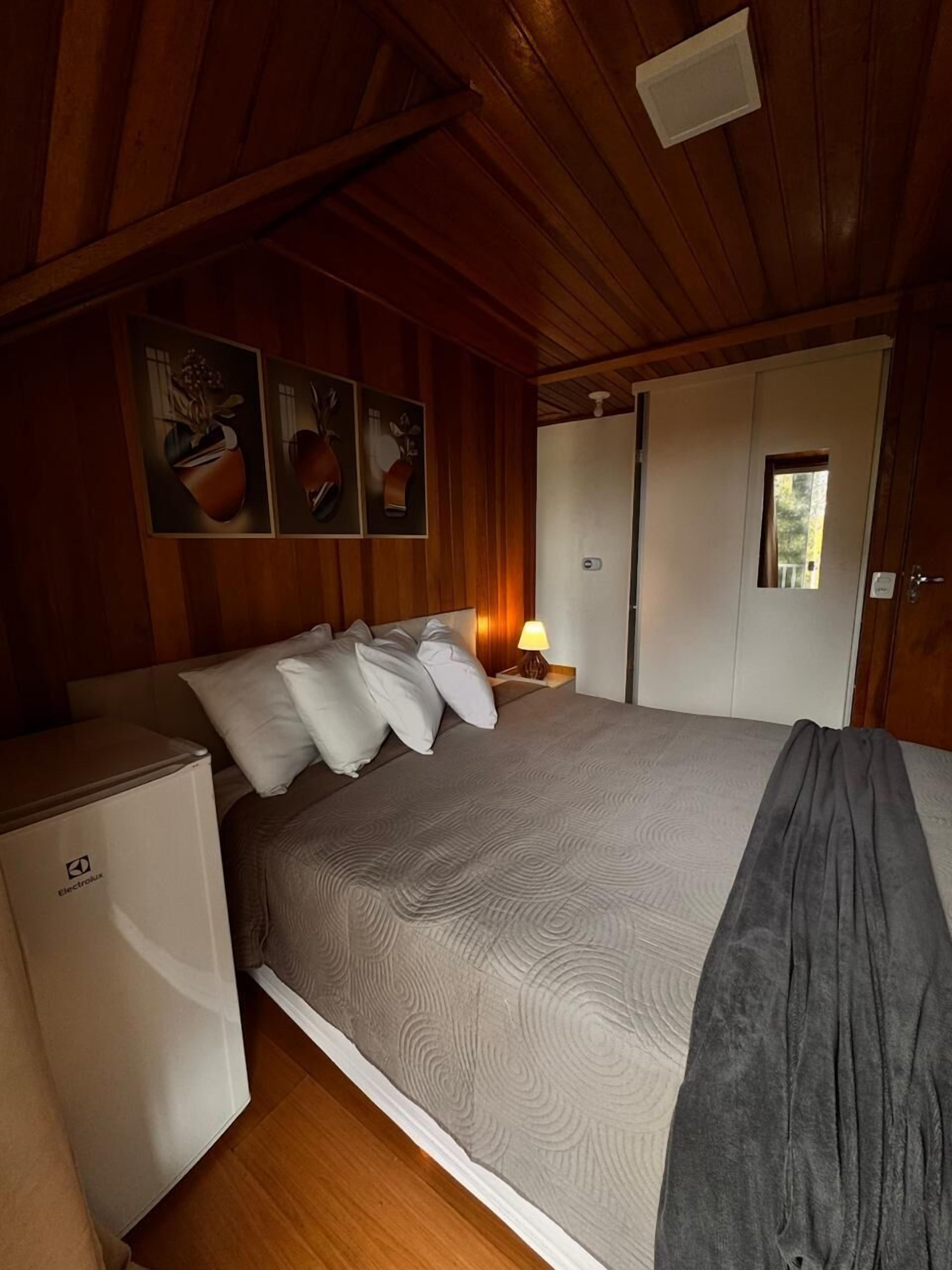 Een slaapkamer met een bed, een nachtkastje met lamp, een koelkast en houten wanden.