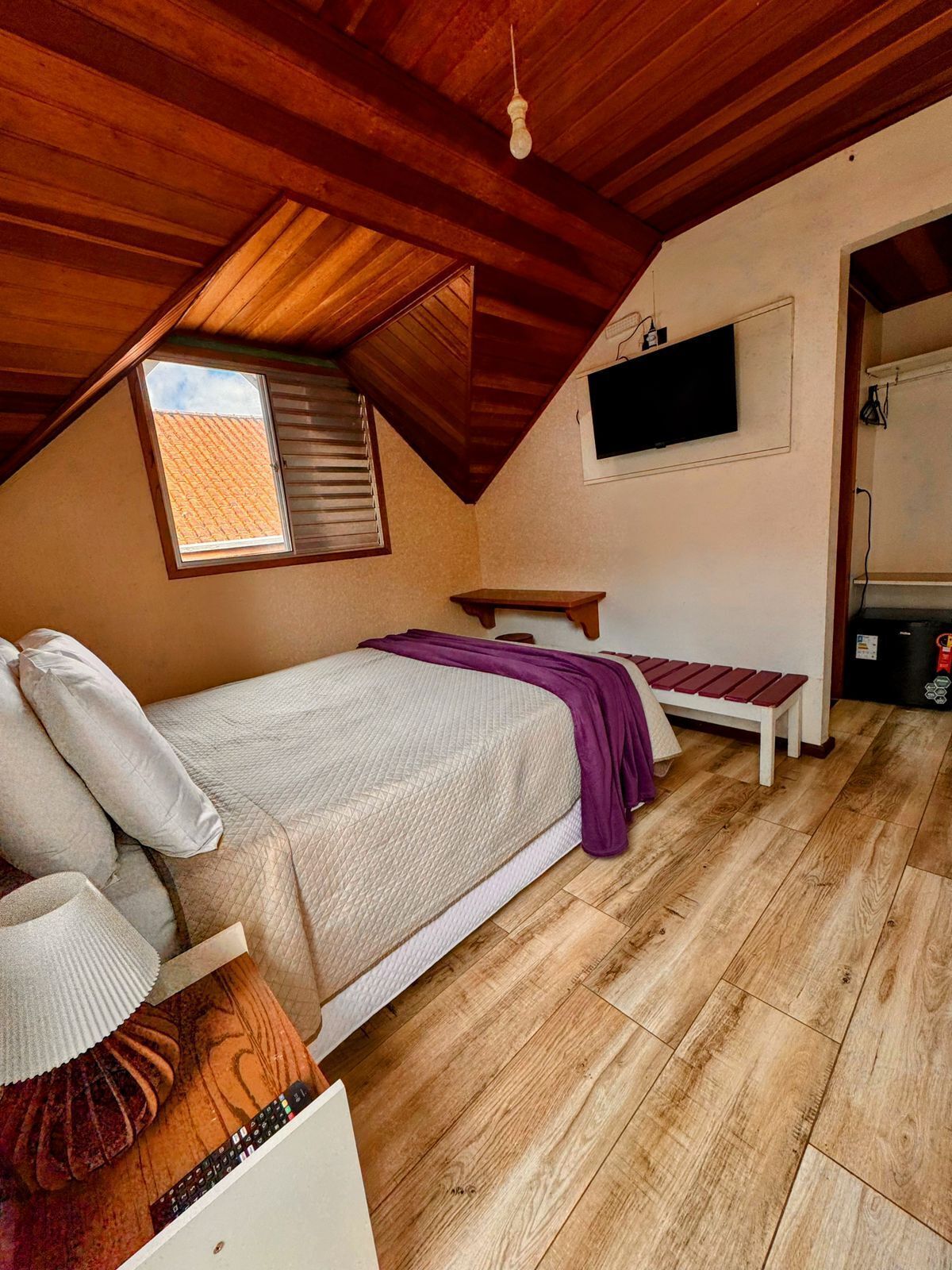 A bedroom with a sloped ceiling, a bed with pillows, a bench, a television, and a window.