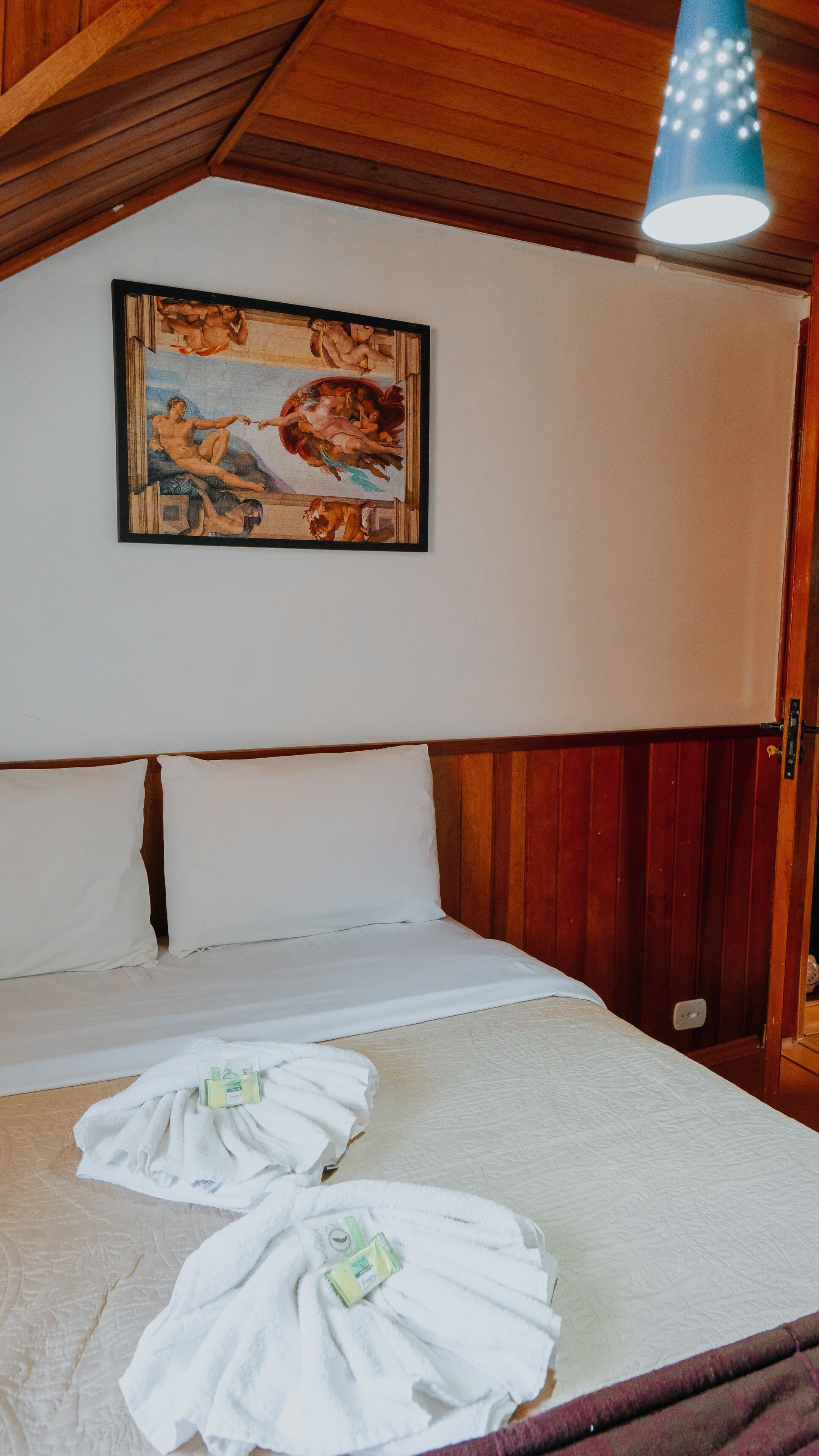 Habitación de hotel con cama, ropa de cama blanca, un cuadro en la pared y una lámpara colgante azul.