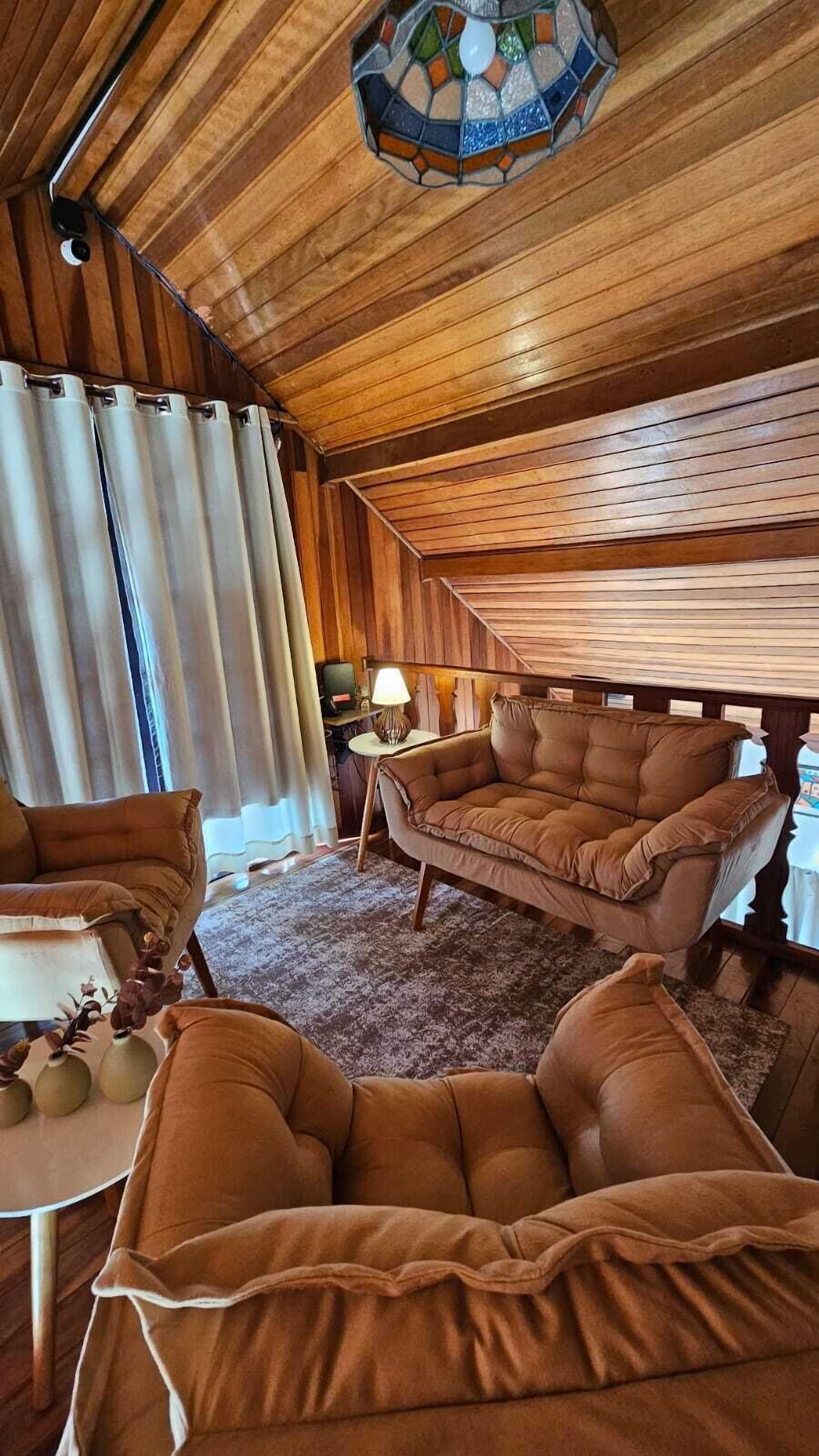 Interior acogedor de una cabaña con paredes de madera, un ventanal con cortinas, un sofá, una silla y una mesita con una lámpara.