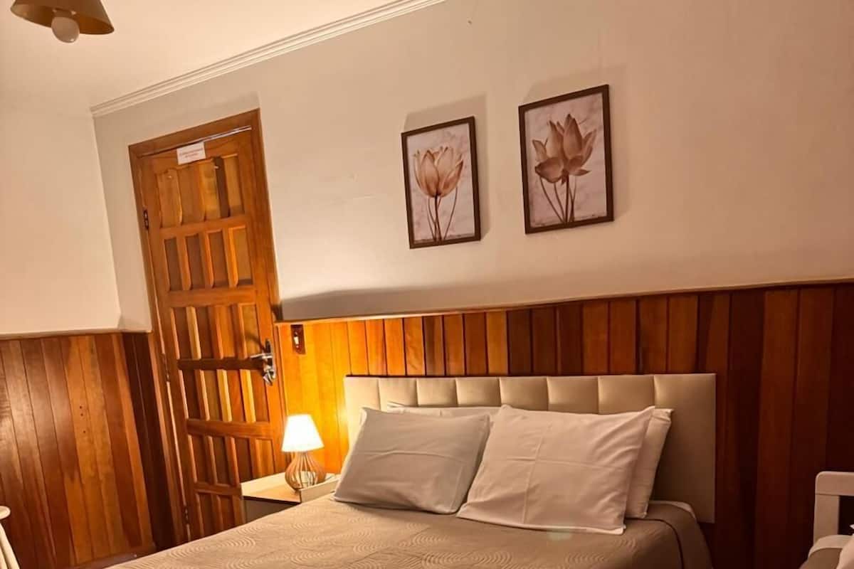 A hotel room with a large bed, a bedside table with a lamp, wooden paneling on the walls, and two framed floral pictures on the wall.