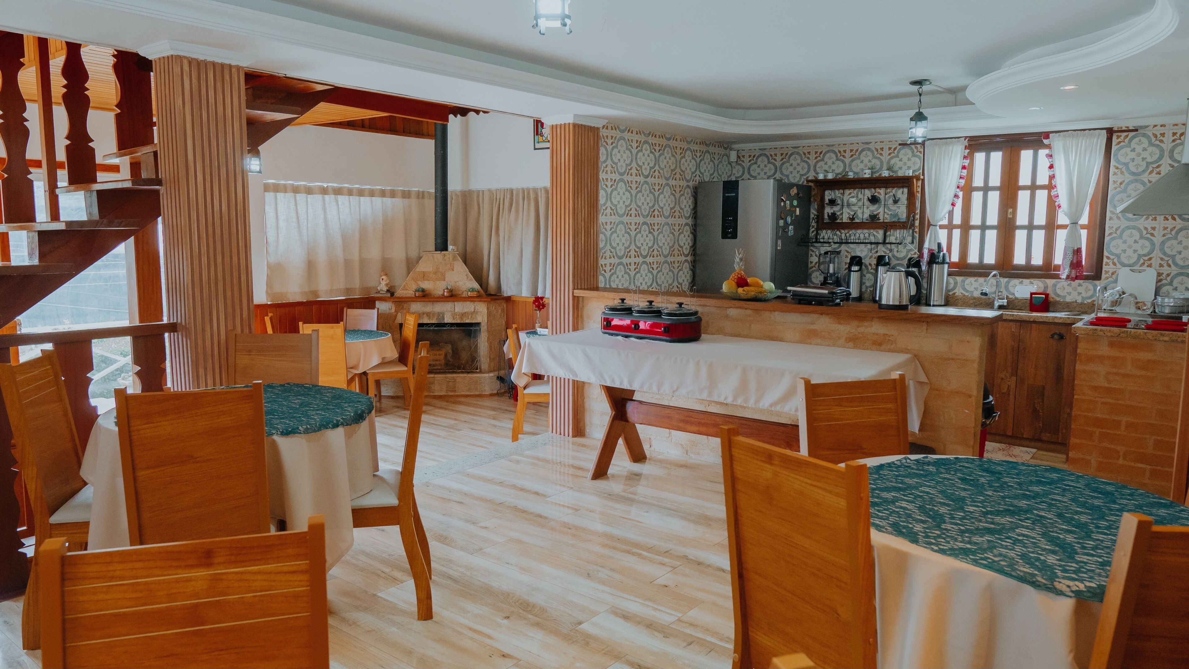 A dining area with wooden chairs and a table, a kitchen with a refrigerator and various appliances, and a staircase leading to an upper level.