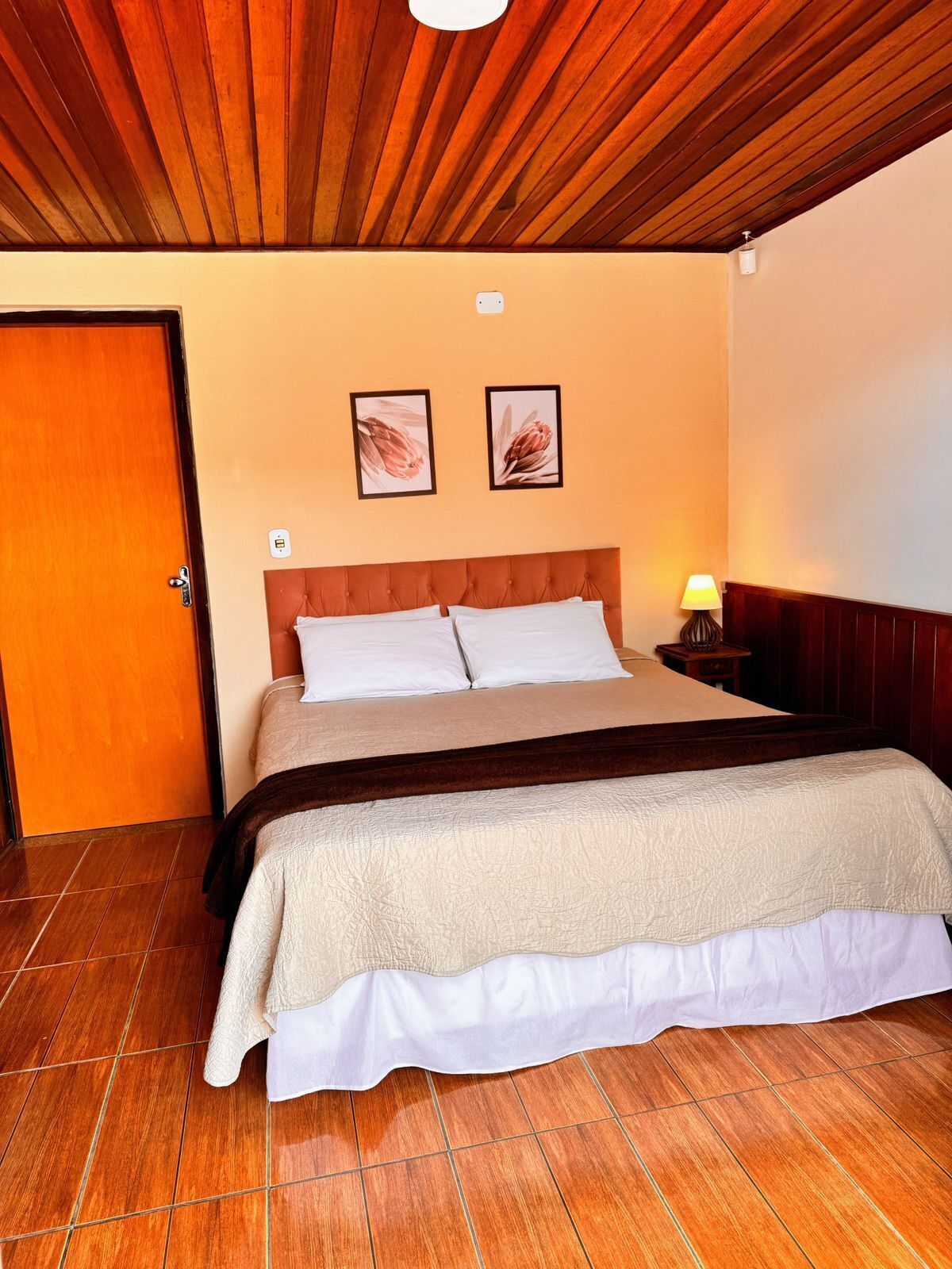 Un dormitorio con techo de madera, una cama con ropa de cama blanca y beige, dos cuadros en la pared y una mesita de noche con lámpara.