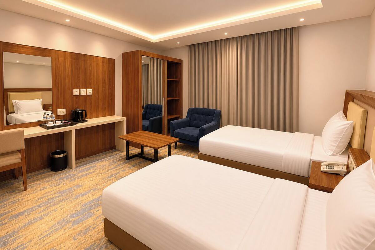 Quarto de hotel com duas camas, uma escrivaninha com cadeira, uma mesinha, um espelho e um guarda-roupa.