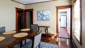 A dining area with a wooden table, chairs, and woven placemats. There is a painting on the wall and a door leading to another room. - Two Bedroom Cutie in Midtown (Omaha)