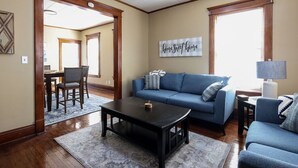 A living room with a blue sofa, a coffee table, and a lamp. There is a dining area in the background with a table and chairs. - Two Bedroom Cutie in Midtown (Omaha)