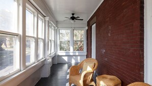 A covered porch with wicker chairs, large windows, and a ceiling fan. - Two Bedroom Cutie in Midtown (Omaha)