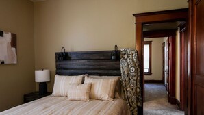 A hotel room with a large bed, a headboard with a dark wood finish, a nightstand with a lamp, and a view of a hallway with wooden doors. - Two Bedroom Cutie in Midtown (Omaha)