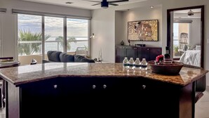 A modern living room with a large window overlooking the sea, a ceiling fan, and a dining area with a marble island. - 5-Star LaJollaReaCondo-Pools&BeachAccess-Rosarito (Rosarito)