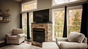 A living room with a stone fireplace, two armchairs, a large flat-screen TV, and a view of a forest through large windows. - Cozy private getaway built for couples to reconnect !!  (West Union)