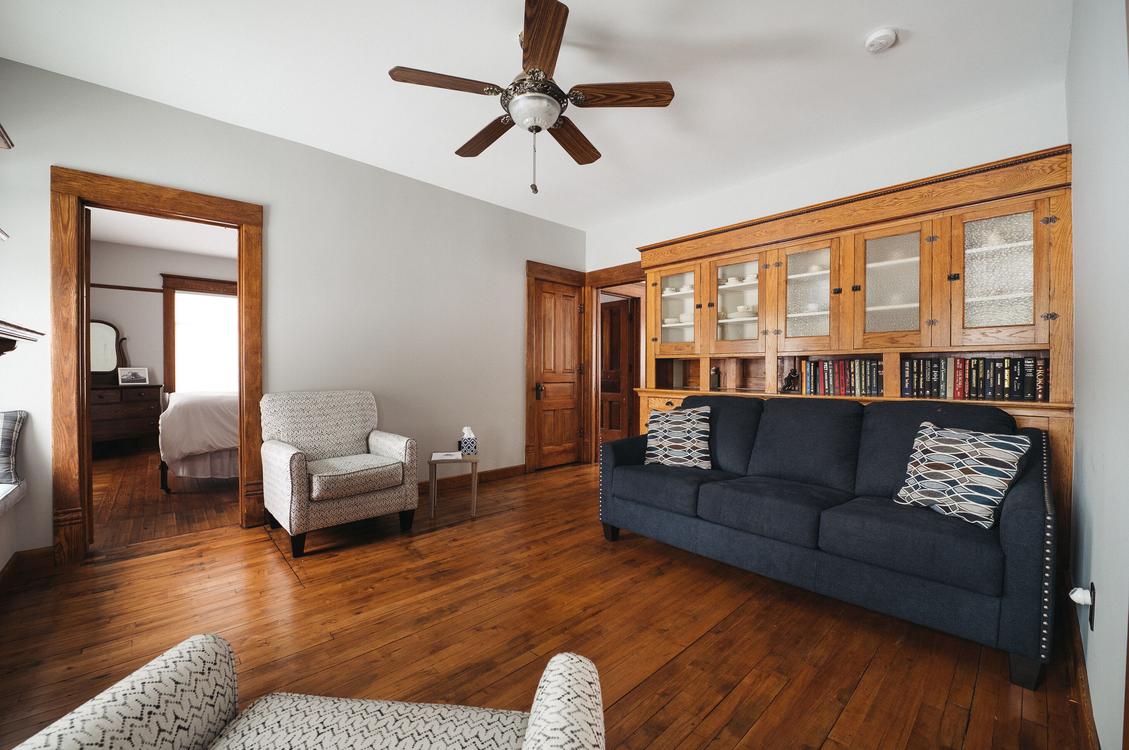 A spacious living room with hardwood flooring, a dark blue sofa, a patterned armchair, and a ceiling fan.
