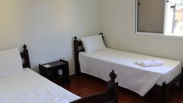 A room with two single beds, each with a wooden headboard and white bedding. There is a small table with a lamp between the beds, and a window with a blind partially drawn.