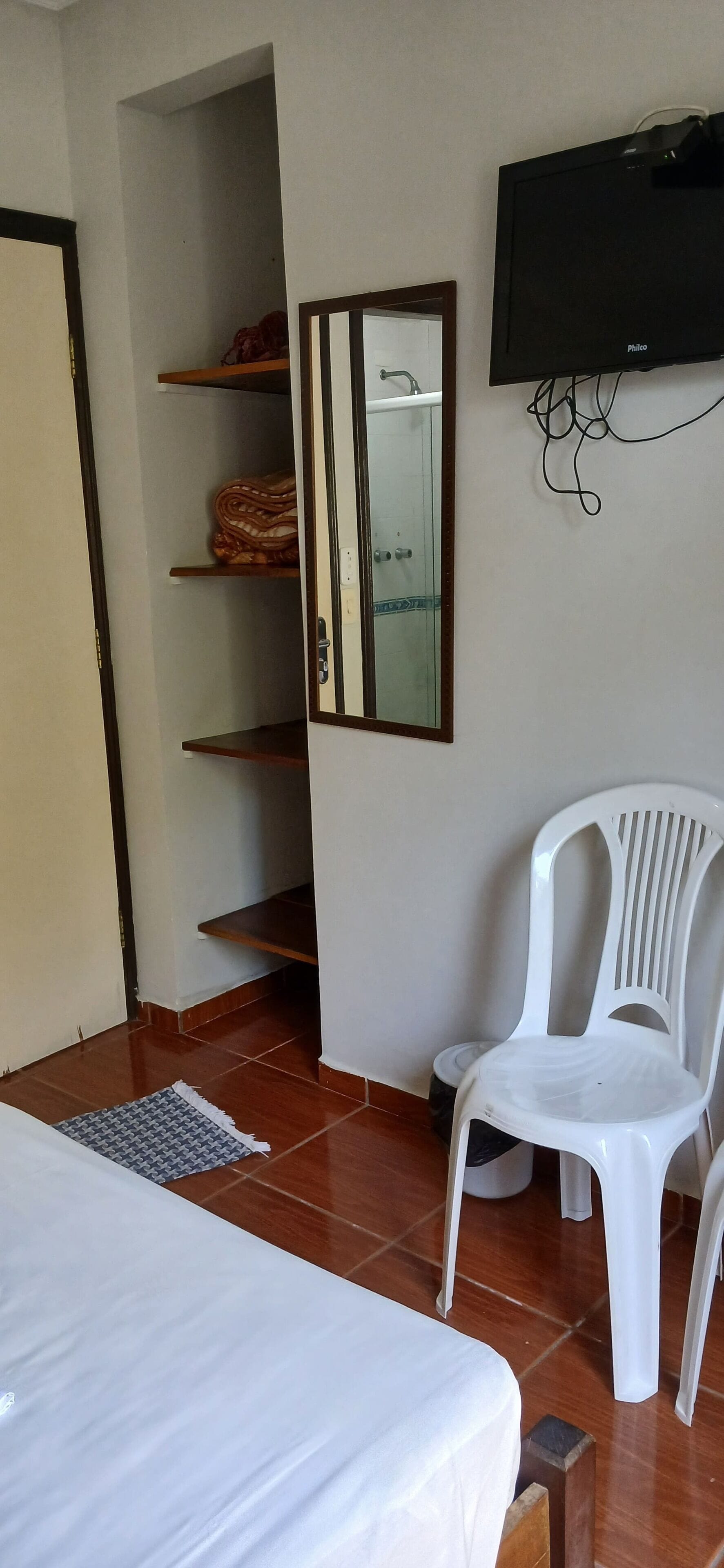Una habitació amb un llit, una cadira de plàstic blanc, una televisió i un mirall.