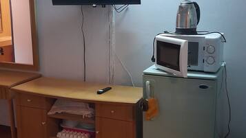 Une chambre avec un téléviseur fixé au mur, un petit réfrigérateur sur lequel est posé un micro-ondes, un bureau en bois avec une télécommande, et un lit recouvert d’une couverture bleue.