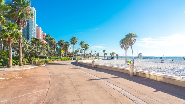 Un paseo marítimo frente a la playa con palmeras, arena y un hotel al fondo.