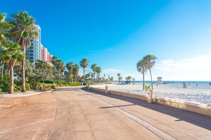 Un paseo marítimo frente a la playa con palmeras, arena y un hotel al fondo.