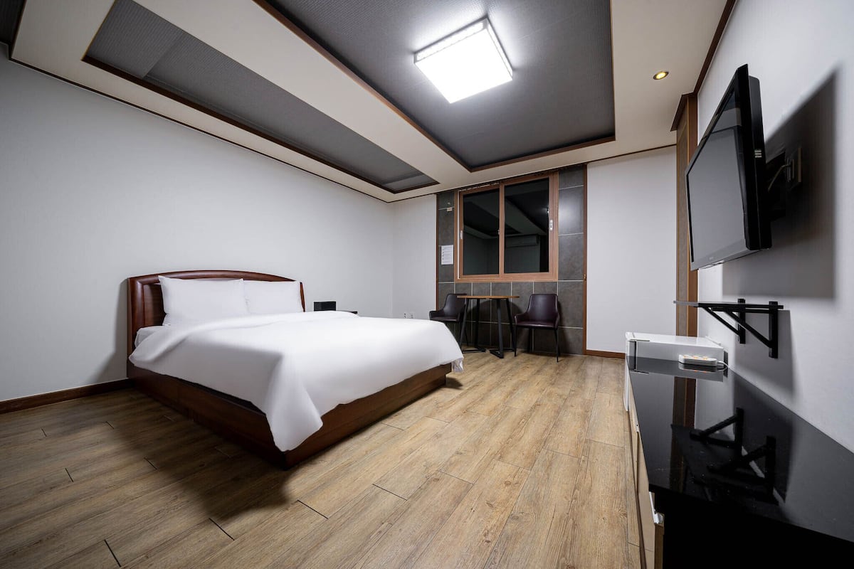A modern hotel room with a wooden bed, a desk with two chairs, a flat-screen TV mounted on the wall, and a large window with a view.