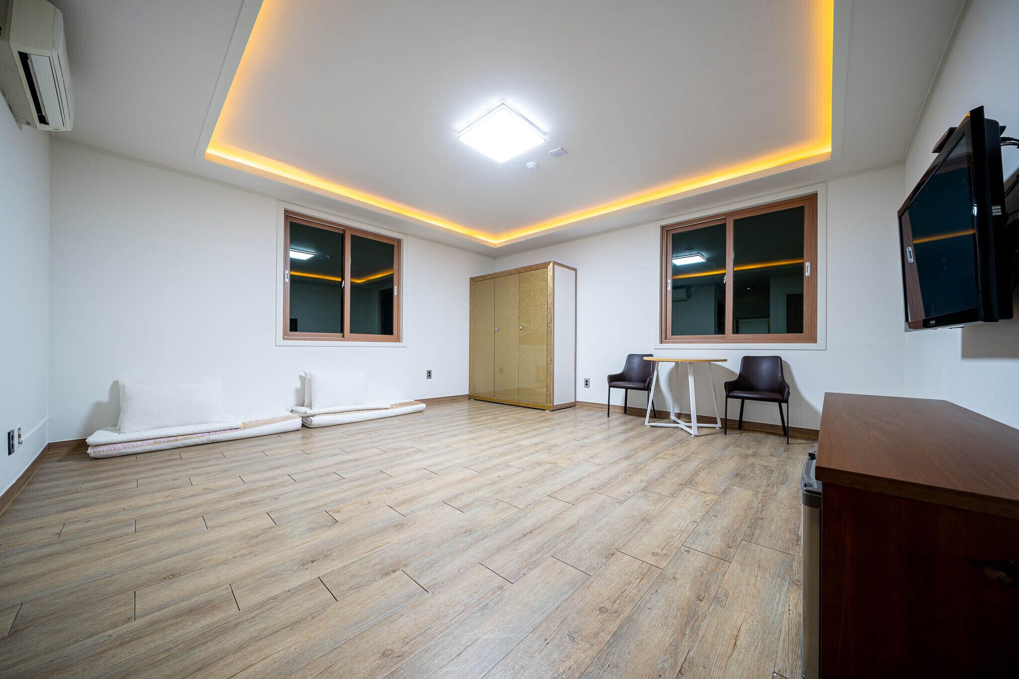 A spacious room with wooden flooring, a large window, a door, a flat-screen TV, and a small table with chairs.