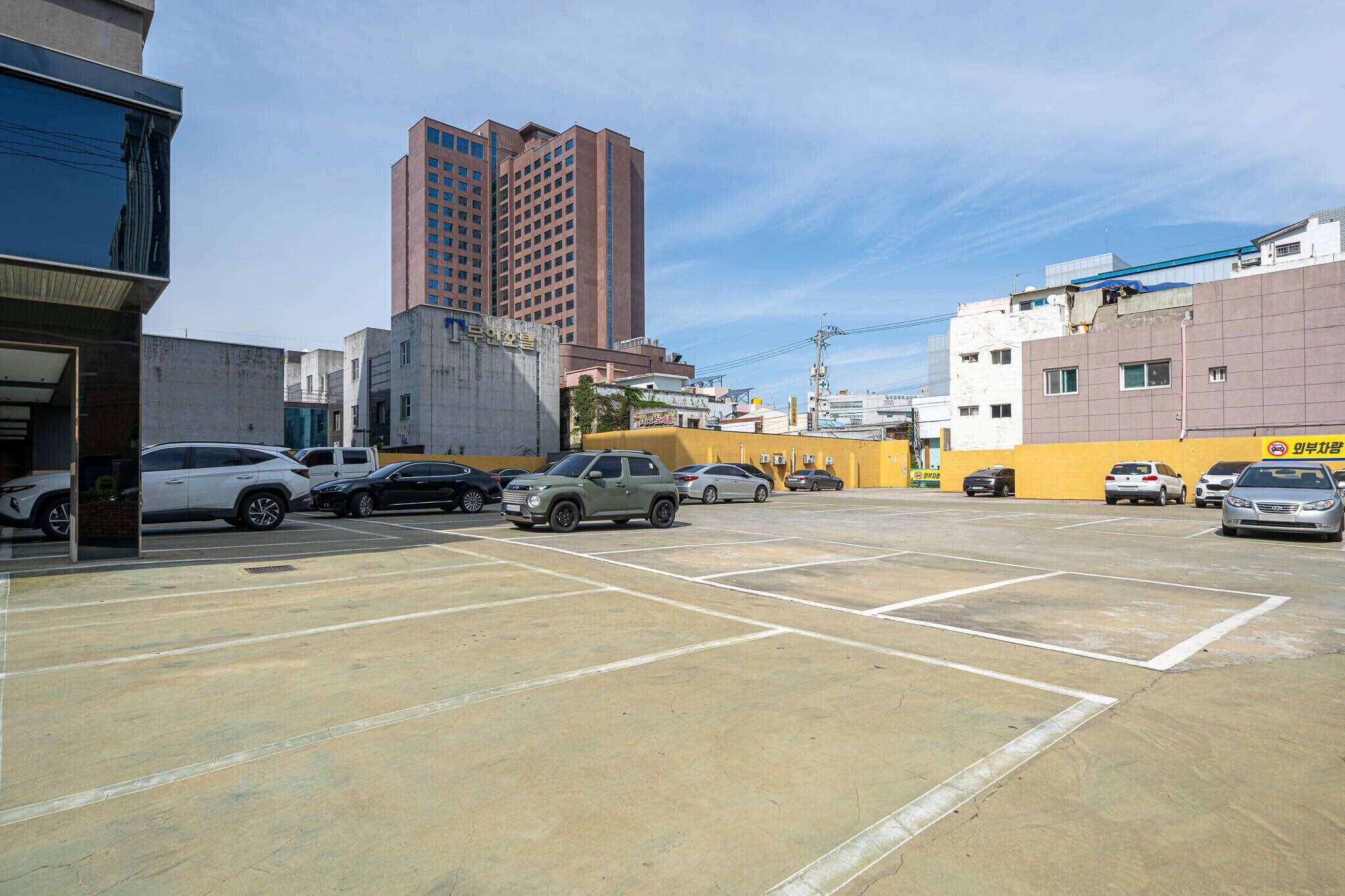 Ein Parkplatz mit mehreren Autos, ein Gebäude mit einer gelben Fassade und ein hohes rotes Gebäude im Hintergrund.