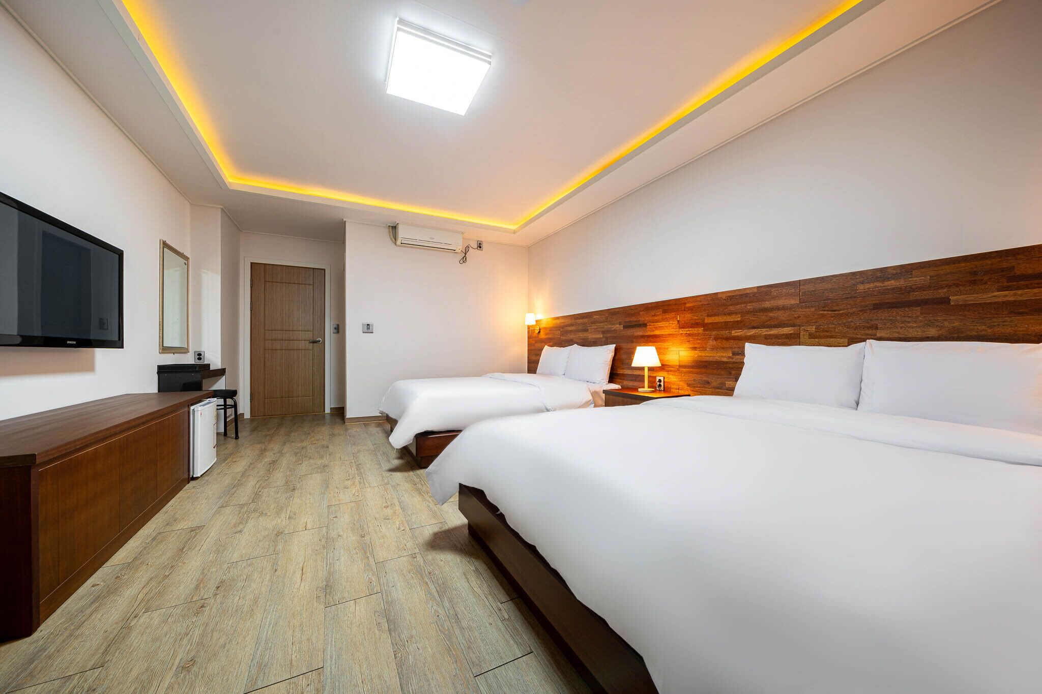 A hotel room with two beds, a wooden headboard, a flat-screen TV, a desk, and a small kitchenette.