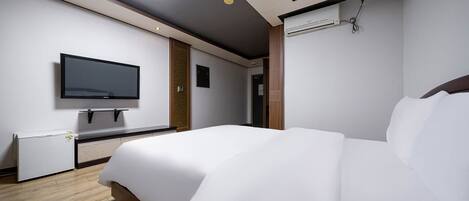 A modern hotel room with a large bed, a flat-screen TV mounted on the wall, a small refrigerator, and a ceiling-mounted air conditioning unit.