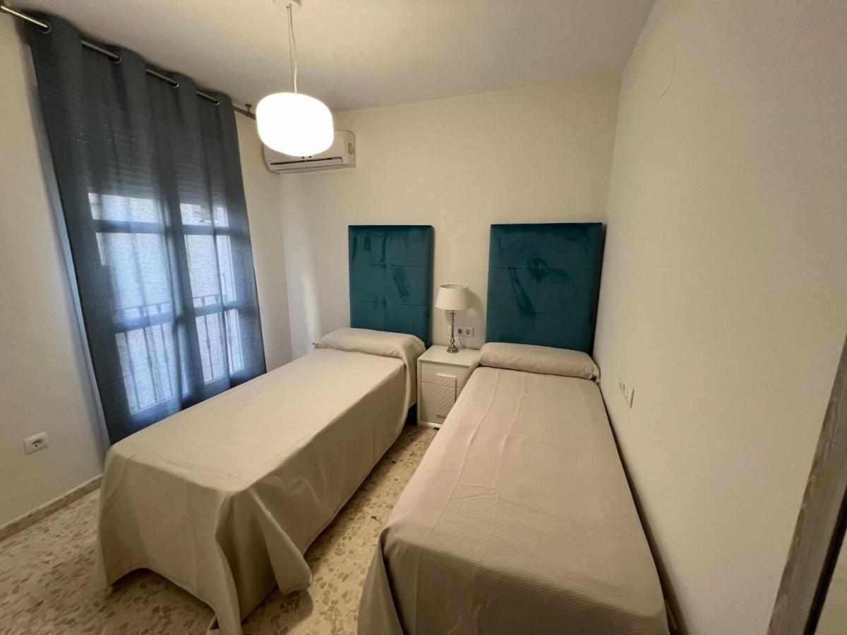 A small, simple room with two beds, a nightstand, a lamp, and a window with curtains.