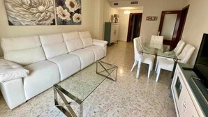 A modern living room with a white sofa, a glass coffee table, and a dining area with white chairs. There are floral paintings on the wall and a chandelier above the seating area. - AREVALO CENTER APARTMENT Free parking (Lucena)